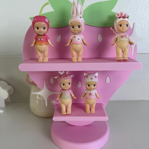 May include: A pink strawberry-shaped shelf displays five small figurines. The figurines have different hats and outfits. The shelf has three levels and a green leaf-shaped top. The figurines are in various poses.