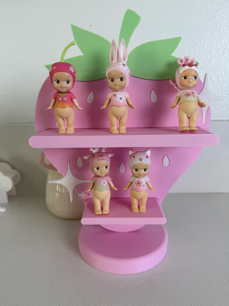 May include: A pink strawberry-shaped shelf displays five small figurines. The figurines have different hats and outfits. The shelf has three levels and a green leaf-shaped top. The figurines are in various poses.