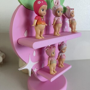 May include: A pink strawberry-shaped shelf with three tiers, holding four small figurines. The figurines are wearing hats and shirts in shades of pink and red. The shelf has a star-shaped accent and green leaves.
