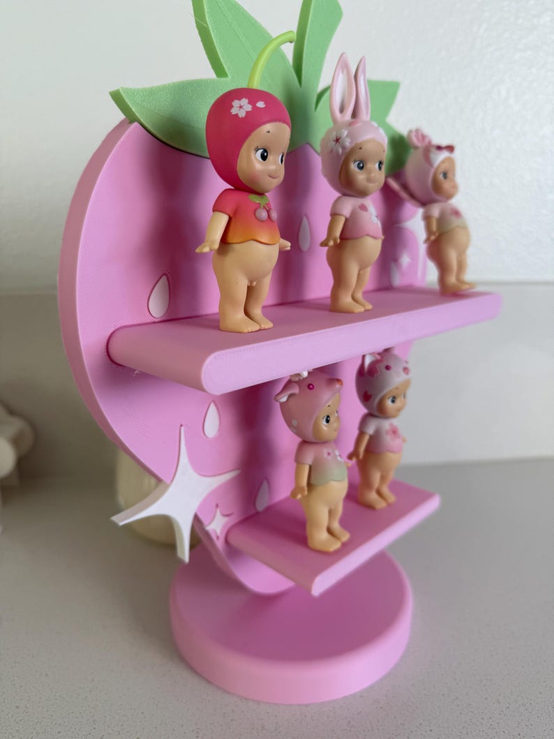 May include: A pink strawberry-shaped shelf with three tiers, holding four small figurines. The figurines are wearing hats and shirts in shades of pink and red. The shelf has a star-shaped accent and green leaves.