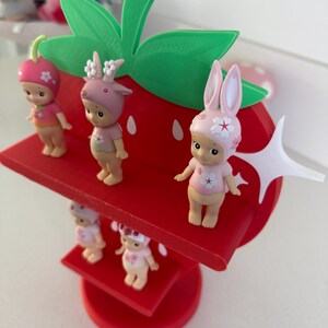 May include: A whimsical red strawberry-shaped display stand with green leaves, holding several small figurines. The figurines are dressed in pink and white outfits, some with animal-themed hats. The stand is a charming decorative piece.