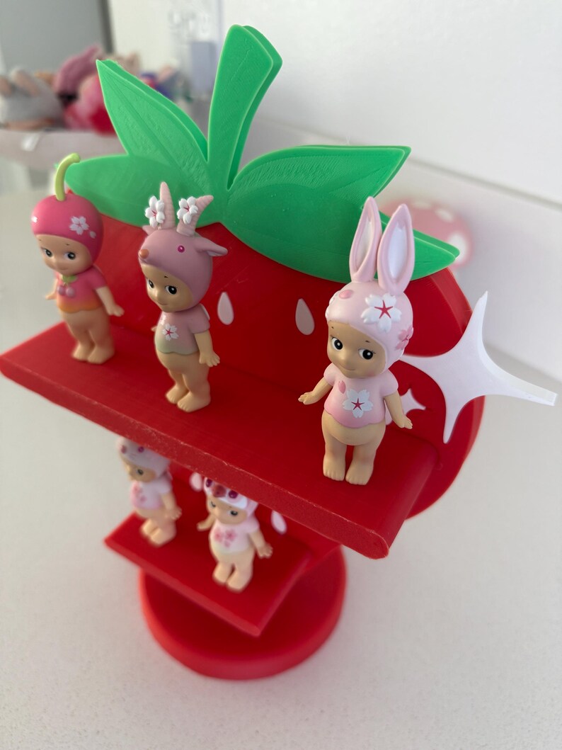 May include: A whimsical red strawberry-shaped display stand with green leaves, holding several small figurines. The figurines are dressed in pink and white outfits, some with animal-themed hats. The stand is a charming decorative piece.