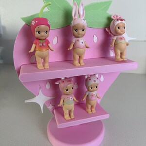 May include: A pink strawberry-shaped shelf with five small figurines. The figurines are wearing hats and tops with floral and fruit designs. The shelf has two levels and a green leaf and stem design at the top. The figurines are in various poses.