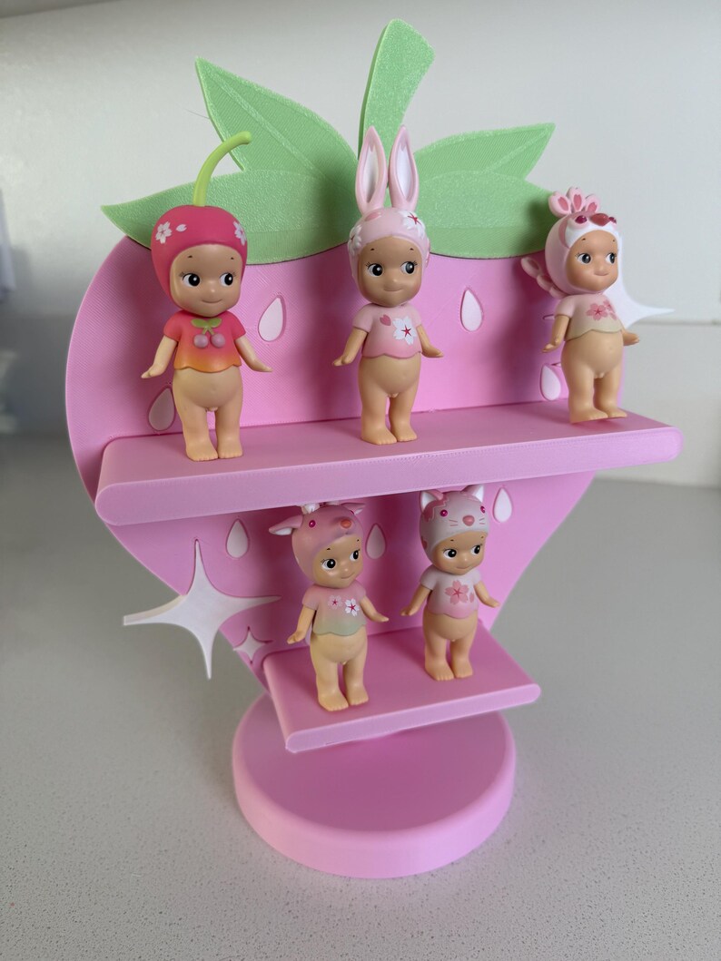 May include: A pink strawberry-shaped shelf with five small figurines. The figurines are wearing hats and tops with floral and fruit designs. The shelf has two levels and a green leaf and stem design at the top. The figurines are in various poses.