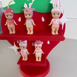 May include: A red strawberry-shaped display stand holding five small figurines. The figurines are nude-colored with pink and white floral accents, wearing hats with animal ears. The stand has green leaves and white starburst details.