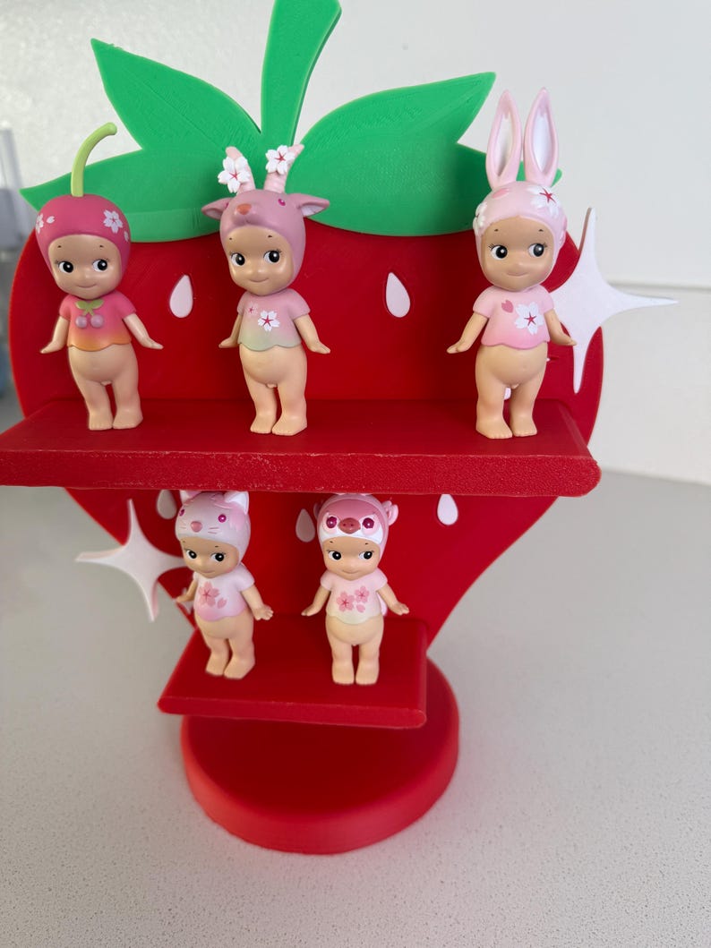 May include: A red strawberry-shaped display stand holding five small figurines. The figurines are nude-colored with pink and white floral accents, wearing hats with animal ears. The stand has green leaves and white starburst details.