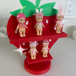 May include: A red strawberry-shaped shelf with green leaves and white seed details, holding five small figurines. The figurines are dressed in pink and beige outfits with floral and animal-themed hats. The shelf is a decorative item.