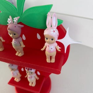 May include: A red strawberry-shaped shelf with green leaves and several small figurines. The figurines are wearing pink outfits and hats with floral and animal-themed designs. The shelf is made of wood and has a tiered design.
