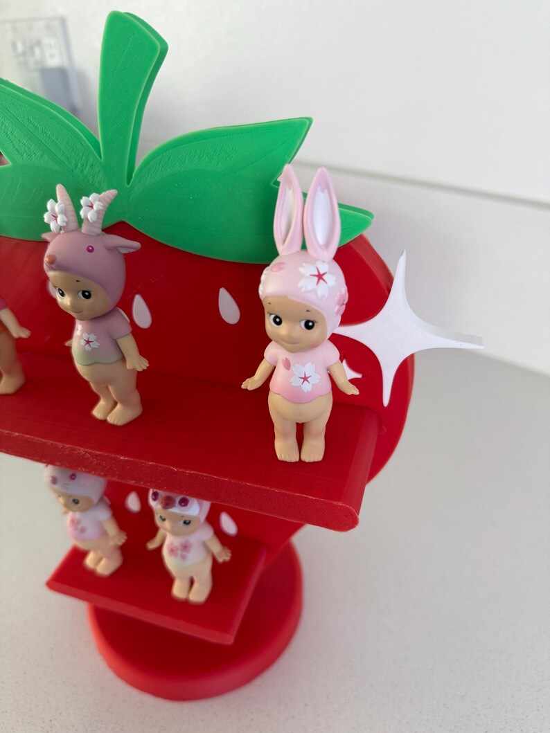 May include: A red strawberry-shaped shelf with green leaves and several small figurines. The figurines are wearing pink outfits and hats with floral and animal-themed designs. The shelf is made of wood and has a tiered design.