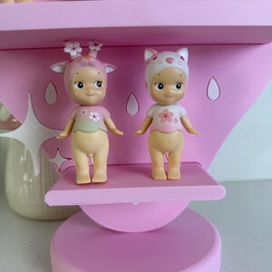May include: Two small, nude-colored figurines with smiling faces and flower-themed outfits stand on a pink shelf. One wears a deer-like headpiece, the other a cat-eared hat. The shelf is part of a larger pink display.