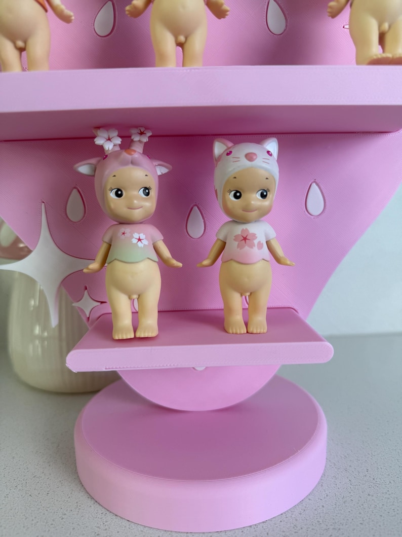May include: Two small, nude-colored figurines with smiling faces and flower-themed outfits stand on a pink shelf. One wears a deer-like headpiece, the other a cat-eared hat. The shelf is part of a larger pink display.