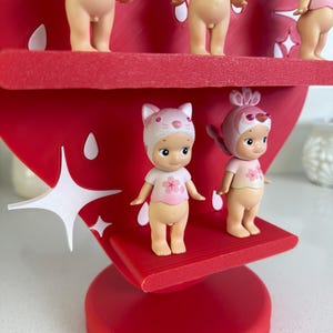 May include: A red strawberry-shaped display stand holding several small figurines. The figurines are nude with pink tops and various head coverings. The stand has white star-shaped accents and is on a white surface.