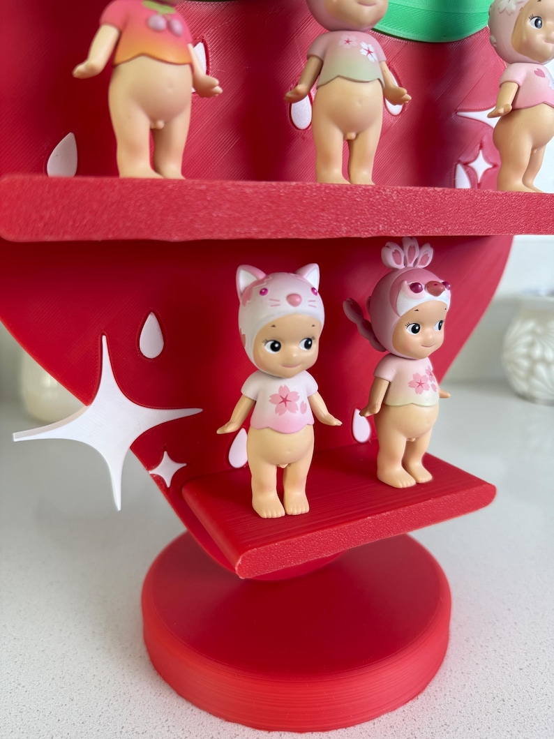 May include: A red strawberry-shaped display stand holding several small figurines. The figurines are nude with pink tops and various head coverings. The stand has white star-shaped accents and is on a white surface.