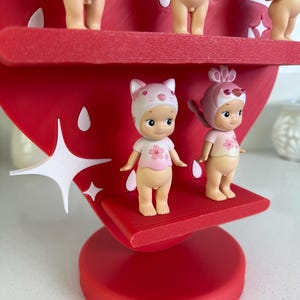 May include: A red strawberry-shaped display stand with multiple small figurines. The figurines are nude with pink tops and hats. The stand has white star-shaped accents and a round base. The figurines are arranged on red shelves.