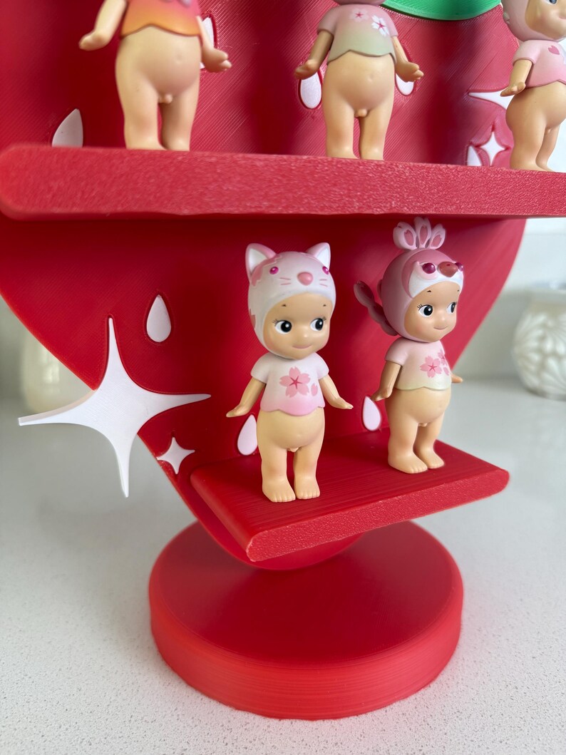 May include: A red strawberry-shaped display stand with multiple small figurines. The figurines are nude with pink tops and hats. The stand has white star-shaped accents and a round base. The figurines are arranged on red shelves.