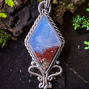 May include: A silver pendant necklace with a diamond-shaped stone. The stone features a blue and red landscape design. The pendant is suspended from a thin silver chain. The pendant has a decorative silver bail and a detailed silver bottom.