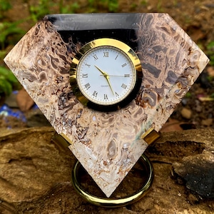 May include: A diamond-shaped desk clock with a gold-tone clock face and Roman numerals. The clock is set in a stone base with brown and cream patterns, and a black top. The clock has a gold-tone stand.