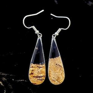May include: A pair of teardrop-shaped earrings with silver hooks. The top half of each earring is black, while the bottom half is a light brown with darker brown veining. The earrings are set against a black background.