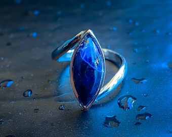 May include: A silver ring with a marquise-shaped, deep blue gemstone. The gemstone is set in a silver band that curves around the finger. The ring is displayed on a dark surface with water droplets, creating a reflective effect.