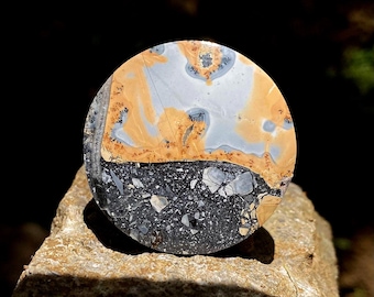 May include: A round, polished stone with a split design. The top half features a blend of light blue, tan, and orange hues, while the bottom half is a speckled black and gray. The stone is set on a rough, tan rock surface.