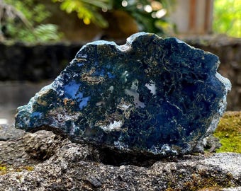 May include: A natural, rough-cut stone displaying a deep blue and green colour scheme. The stone features complex patterns and a polished look, with lighter blue and white sections. It rests on a textured rock surface.