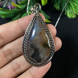 Può includere: Un ciondolo d'argento con una pietra di agata a forma di lacrima. La pietra presenta una miscela di colori bianco, grigio e nero con un motivo a spirale. Il ciondolo è bordato da un bordo d'argento a forma di corda.