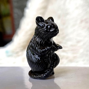 May include: A glossy black squirrel figurine, sculpted in a seated pose. The sculpture has detailed fur texture and a smooth, reflective surface. The squirrel is sitting upright, with its paws resting on its belly. The figurine is a decorative item.