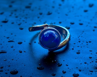 May include: A silver ring with a large, round, deep blue gemstone. The ring has a simple, elegant design with a curved band that wraps around the finger. The gemstone is set in a bezel setting, and the ring is displayed on a dark blue surface.