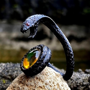 May include: A black snake ring with a detailed, textured design. The snake's head is raised, with a red gemstone eye and open mouth. The ring features an oval-shaped, amber-colored gemstone. The ring is set on a natural stone surface.