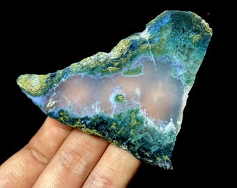 May include: A slice of agate stone with a unique pattern. The stone features a mix of colors, including blue, green, and gold, with a translucent center. The agate is held in a hand against a black background.