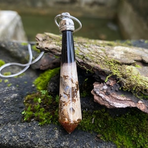 May include: A pendant necklace featuring a polished, elongated gemstone. The top portion is black, transitioning to a beige and brown patterned center, and ending with a reddish-brown point. The pendant is suspended from a silver-colored chain.