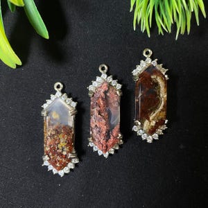 May include: Three silver pendant charms with a red, brown, and white agate stone set in a silver bezel with clear rhinestones around the edge.