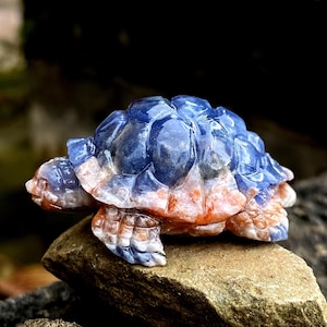 May include: A carved stone turtle figurine with a blue and orange color scheme. The turtle's shell is a deep blue, while its body and legs are a mix of orange and white. The figurine is resting on a natural stone.