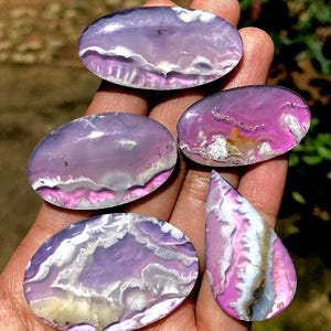 May include: Five polished agate stones in shades of purple, pink, white, and yellow are displayed. The stones have a smooth, glossy surface with unique patterns resembling landscapes. One stone is teardrop-shaped, while the others are oval.