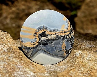 Puede incluir: Una piedra redonda y pulida con un patrón único. La piedra presenta una mezcla de colores, incluyendo azul claro, beige, negro y gris. El diseño se asemeja a un paisaje con detalles intrincados. La piedra está colocada sobre un fondo natural y terroso.