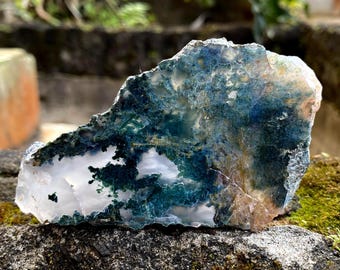 May include: A raw, natural agate stone with a mix of colors. The stone features areas of translucent white, deep green, and blue, with hints of tan. The rough edges and unique patterns give it a natural, organic appearance.
