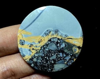 Puede incluir: Una piedra pulida y redonda con un diseño de paisaje. La piedra presenta un cielo azul claro, una cordillera amarilla y negra y un primer plano gris oscuro y negro. La piedra se sostiene en una mano.