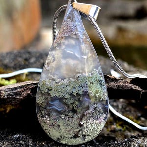 Op de afbeelding: Een druppelvormige hanger met een zilveren ketting. De hanger heeft een heldere, doorschijnende steen met groene en paarse insluitsels, die op een landschap lijken. De hanger is opgehangen aan een zilveren oogje.