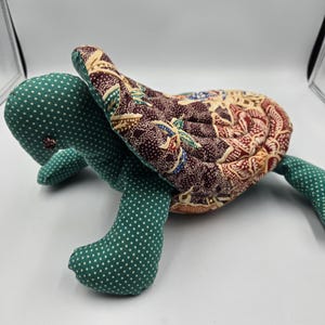 May include: A handmade stuffed turtle toy. The turtle's head and limbs are green with white polka dots. The shell is patterned with a mix of brown, beige, and red floral designs. The toy is soft and plush.
