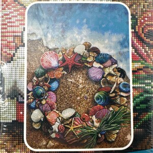 May include: A colorful image of a seashell wreath on a sandy beach with a blue ocean background. The wreath is composed of various seashells, including red, pink, and blue shells, and a red starfish. The image has a rectangular shape with rounded corners.