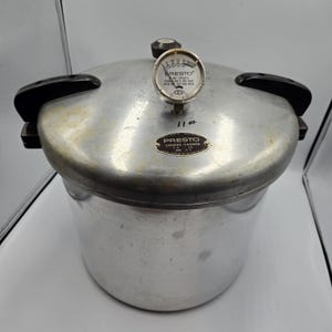 May include: A silver-colored Presto pressure cooker with a gauge and black handles. The gauge reads "Presto" and "Cooker-Canner". The pressure cooker is a vintage kitchen appliance.