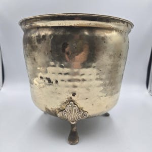 May include: A tarnished, hammered metal planter with a rounded body and three decorative feet. The planter has a wide rim and a slightly textured surface. The feet have an ornate, stylized design.