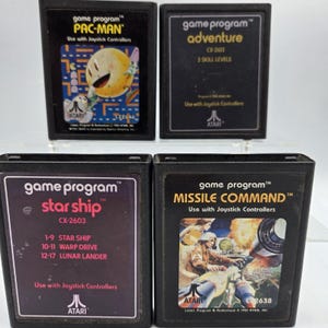 May include: Four vintage Atari game cartridges: Pac-Man, Adventure, Starship, and Missile Command. Each black cartridge features game titles and graphics, with the Atari logo. The Pac-Man cartridge has a yellow character. The games are designed for use with joystick controllers.