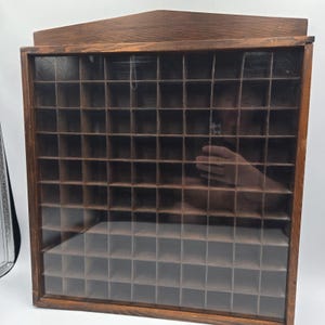 May include: A square wooden display case with a dark brown finish, featuring a grid of numerous small, open compartments. The front of the cabinet is covered by a clear glass panel, and the top edge has a slightly peaked, angular wooden design.