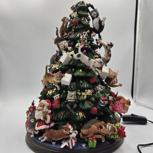 May include: A whimsical Christmas tree figurine featuring numerous cats and dogs climbing and playing among ornaments and gifts. The tree is decorated with red ornaments, candy canes, and lights, with Santa Claus at the base. The figurine is on a dark blue base.