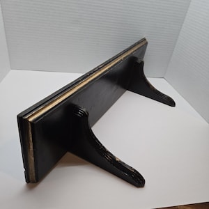 May include: A black wooden shelf with two decorative supports. The shelf has a gold trim along the top edge. The supports are curved and add a vintage aesthetic. The shelf is designed to be mounted on a wall.