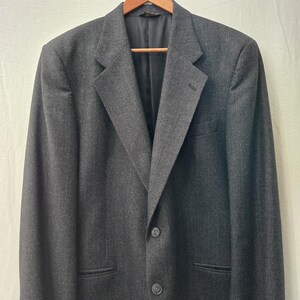 May include: A charcoal gray blazer with a two-button closure, notched lapels, and a chest pocket. The blazer is hanging on a wooden hanger against a neutral background. The jacket has a classic, tailored design.