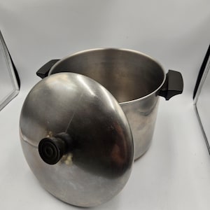 May include: A stainless steel cooking pot with a lid. The pot has two black handles and a lid with a black knob. The lid is partially open, revealing the inside of the pot.