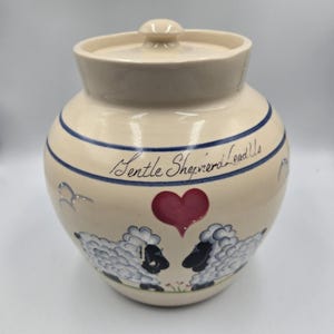 May include: Cream-colored ceramic jar with a lid, featuring blue stripes and a hand-painted design. The jar has two sheep facing each other with a red heart above them. The text "Gentle Shepherds Lead Us" is written in cursive.