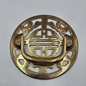 May include: A brass drawer pull with a circular design. The pull features an openwork pattern with a central geometric design and a curved handle. The brass has a warm, aged appearance, suggesting vintage or antique origins.